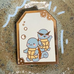 May include: A gold-edged tag featuring two cartoon turtles in blue and yellow. The turtles have pink cheeks and are set against a cream background with bubbles and decorative elements. The tag is on a textured surface.