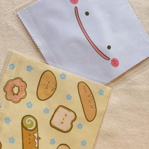May include: Two small, square, clear plastic pouches. One is white with a smiling face design. The other is yellow with illustrations of baked goods, including a donut, bread, and a roll, with small blue flower accents.