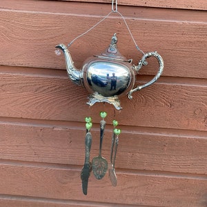 Puede incluir: Un carillón de tetera de plata con cuentas verdes en forma de corazón y cucharas de plata colgando.