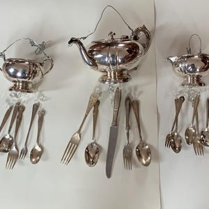 Puede incluir: Tres juegos de té de plata con cubiertos colgantes. Los juegos incluyen una tetera, una crema y un azucarero. Cada juego está suspendido con cuentas y alambre transparentes, con tenedores, cucharas y un cuchillo colgando debajo.