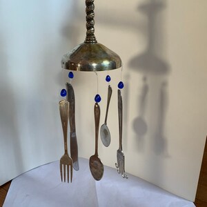 May include: A distinctive wind chime made from recycled silverware. The chime has a silver-coloured base with a twisted stem, suspending forks, spoons, and a knife. Blue teardrop-shaped beads are interspersed between the utensils, adding colour to the design.