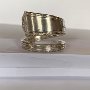 Puede incluir: Un anillo de cuchara de plata, hecho doblando una cuchara en un círculo. El anillo tiene un borde decorativo y está hecho de una cuchara vintage.