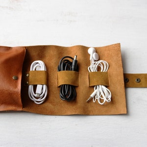 May include: A brown leather cord organizer with three compartments for holding cords and headphones. The organizer has a flap that closes with a snap and a strap with snaps to secure it.