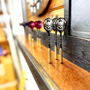 May include: A set of darts with black and white skull and crossbones design flights, gold barrels, and black shafts. The darts are arranged on a wooden surface, with a dartboard and a dark wooden wall in the background.