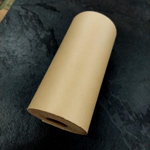 May include: A roll of brown kraft paper. The paper is tightly wound into a cylindrical shape. The end of the roll reveals the inner core. The paper has a smooth, matte finish. Ideal for wrapping, crafting, or packaging.