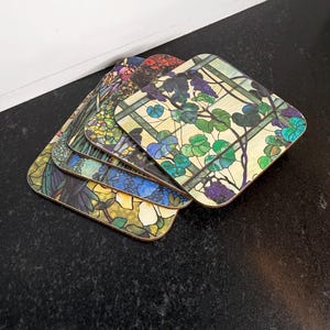 May include: A stack of four square coasters with colorful floral and botanical designs. Each coaster features a different stained-glass style pattern with shades of green, blue, purple, and yellow. The coasters are made of a durable material and have a smooth, glossy finish.