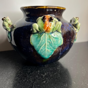 May include: A dark blue and brown glazed ceramic vase featuring three-dimensional sculpted frogs perched on green lily pads. The vase has a rounded body with a wide opening and a glossy finish, showcasing intricate details on the frog figures.
