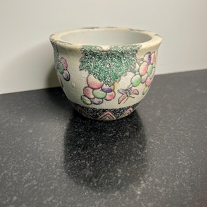 May include: A small white ceramic planter with a grape design. The planter has green leaves, pink and purple grapes, and a butterfly.