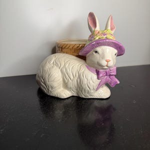 May include: A ceramic figurine of a white rabbit wearing a purple bonnet with a floral pattern and a matching purple bow around its neck. The rabbit is positioned in front of a small, brown, woven-textured basket container for holding items.