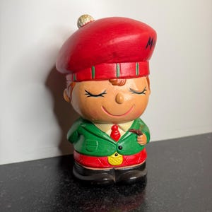May include: A ceramic figurine of a smiling boy wearing a red beret with the letter "M", a green jacket, red tie, and black pants. The figurine is holding a pipe and has closed eyes.