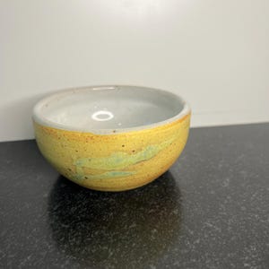 May include: A small, yellow ceramic bowl with a white interior and a light blue glaze. The bowl has a speckled, rustic look.