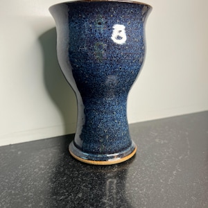 May include: A handmade ceramic vase with a speckled, dark blue glaze. The vase has a flared rim, a slender neck, and a wide base. The base has a light brown rim. The vase is approximately 6 inches tall.
