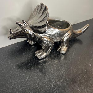 May include: A silver-colored ceramic Triceratops planter. The dinosaur has three horns, a frill, and a tail. The planter has a large opening on its back for plants or other decorative items. The surface has a shiny, metallic finish.