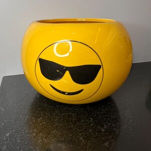 May include: A yellow ceramic bowl shaped like a smiley face with black sunglasses. The bowl has a glossy finish.
