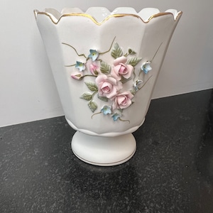 May include: White ceramic vase with a scalloped rim and a gold trim. The vase features a raised floral design with pink roses and blue flowers.