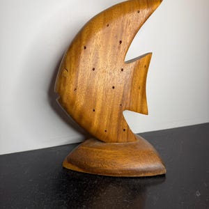 May include: A wooden fish sculpture with a curved body and fins. The sculpture is made of a light brown wood with a visible grain and small drilled holes. It sits on a rounded base, and the background is a plain white wall.