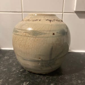 Peut inclure: Un vase rond en céramique blanc cassé avec un glaçage craquelé et de subtiles stries bleu-vert. Le vase a un bord légèrement texturé et un corps bulbeux. C'est un objet décoratif pour la décoration intérieure.