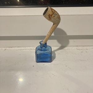 May include: A vintage smoking pipe with a curved stem, placed in a small, square, blue glass inkwell. The pipe has a light brown, aged look. The inkwell is empty and sits on a white surface.