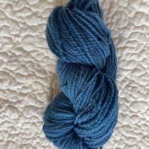 Peut inclure: Une pelote de laine bleu indigo, bien enroulée et texturée. La laine est d'un bleu profond et saturé, idéale pour les projets de tricot ou de crochet. La laine est présentée sur un fond blanc matelassé.