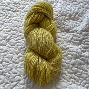 May include: A skein of yellow-green yarn, tightly wound and textured, rests on a white quilted surface. The yarn appears soft and ready for knitting or crocheting projects. The color is a vibrant, natural hue.