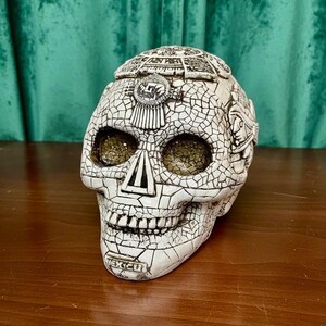 May include: A white skull sculpture featuring a black crackle finish and intricate relief carvings. The top displays a circular calendar design, while the forehead shows a central emblem. The word MEXICO is embossed on the chin area.