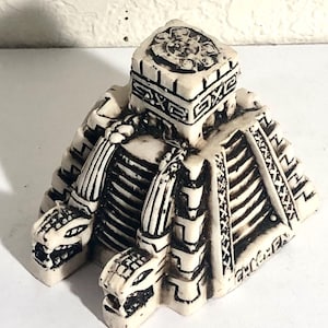 May include: Ivory-colored, detailed figurine depicting a Mesoamerican pyramid with serpent heads. The sculpture features intricate carvings and a square top with a circular design. The pyramid has multiple levels and a textured surface.