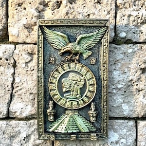 May include: A rectangular wall plaque with a relief design featuring an eagle with outstretched wings, a circular emblem with a figure, and a pyramid. The plaque has a bronze and green color scheme, with intricate details and a textured surface.
