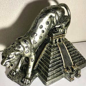 May include: A silver jaguar statue perched atop a silver pyramid with gold accents. The jaguar is depicted in a crouching position with its head turned to the side. The pyramid has a series of steps leading up to the top.