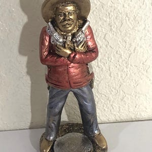 May include: A figurine of a man in a sombrero and red jacket, holding two silver pistols. The figure has a gold-colored hat and boots, with blue pants. The base is a gold-colored circle.
