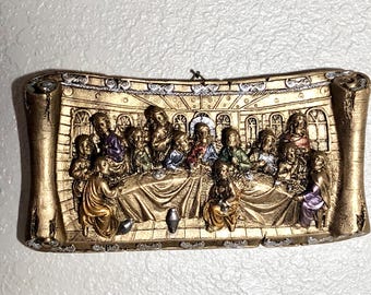 Könnte beinhalten: Eine goldfarbene Wandplakette, die das Abendmahl darstellt. Die Szene spielt in einem Raum mit Bogenfenstern und einem Tisch, der mit einer goldenen Tischdecke bedeckt ist. Die Figuren sind in Relief gearbeitet und in gedeckten Farben bemalt.