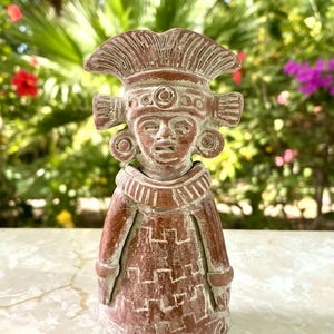 May include: A terracotta-colored clay figurine depicting a figure wearing an elaborate headdress, circular ear ornaments, and a wide collar. The figure is dressed in a long garment featuring a repeating geometric pattern etched into the surface.