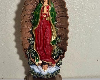 May include: A religious figurine of the Virgin Mary, depicted in a red dress and green cloak, with hands clasped in prayer. The statue is set against a brown halo-shaped backdrop and rests on a copper-colored base. An angel is at the bottom.