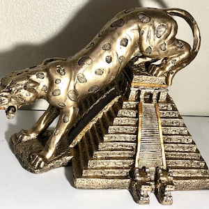 May include: A gold-colored jaguar statue perched atop a miniature replica of a Mayan pyramid. The jaguar is depicted in a crouching position, with its head turned to the side and its mouth slightly open. The pyramid is detailed with intricate carvings and steps.