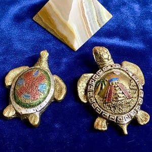 May include: Two gold-toned metal turtle ornaments. The left turtle features a colorful circular Aztec calendar design on its shell. The right turtle displays a raised relief of a pyramid and palm tree on its shell, surrounded by a geometric border.