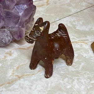 May include: A small, dark brown, hand-sculpted clay figurine of a four-legged animal with pointed ears and a short tail. The figure features light-colored, incised markings on its face and back, and it appears to be holding a small object in its mouth.