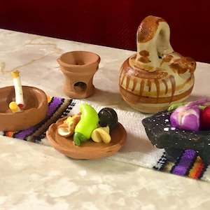 May include: Miniature clay pottery set with a small ashtray holding a cigarette, a small vase, a water jug, a plate with food, and a mortar and pestle. The set includes a colorful woven mat.
