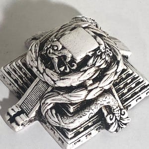 May include: White ceramic figurine of a pyramid with a snake wrapped around it. The snake has a detailed design and is curled around the top of the pyramid. The pyramid has a square base and a pointed top.
