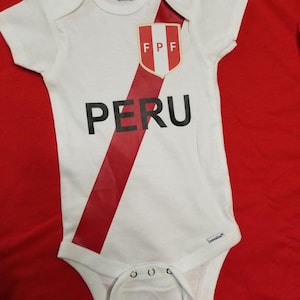 May include: White baby onesie with a red diagonal stripe and the words "PERU" in black. The onesie also features a red and white crest with the letters "FPF" in gold.