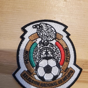 May include: A circular embroidered patch featuring a black and white soccer ball at the bottom, an eagle at the top, and green and red accents. The text reads PASION, ORGULLO, and SELECCION NACIONAL DE MEXICO in black lettering on a gold background.