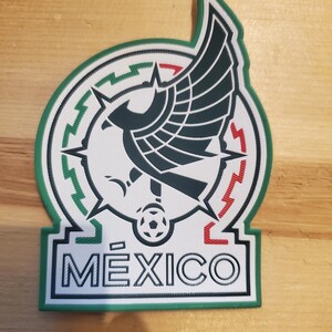 May include: A rubberized patch featuring a stylized black eagle emblem centered within a circular frame. The border incorporates green, white, and red geometric patterns. Below the emblem, a soccer ball sits above the text MEXICO in black letters.