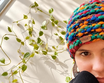 May include: A chunky knit beanie featuring a vibrant, multi-colored pattern of blue, orange, green, and red yarn. A small, rectangular brown leather tag is attached to the front of the beanie with the word Hygge embossed in black lettering.