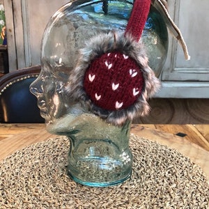 Peut inclure: Des oreillettes en tricot rouge avec des cœurs blancs et une bordure en fausse fourrure. Les oreillettes sont présentées sur une tête de mannequin.