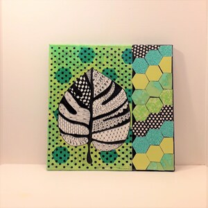 May include: A black and white leaf with a detailed pattern is painted on a green canvas with a green and black polka dot pattern. The right side of the canvas has a green, yellow, and black geometric pattern.