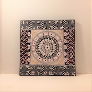 May include: A square wall art piece with a pink and gray mandala design in the center. The mandala is surrounded by a border of pink and gray geometric shapes. The background is a black and white geometric pattern.
