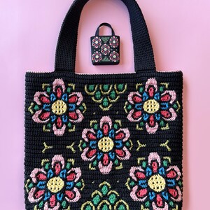 May include: Black crochet tote bag and matching mini bag, both adorned with colorful floral designs. The large tote features large, detailed flowers in pink, yellow, blue, and green. The smaller bag mirrors the design, offering a coordinated set.