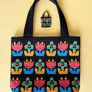 Könnte beinhalten: Eine marineblaue Häkeltasche und eine passende kleine Tasche. Beide Taschen zeigen ein sich wiederholendes Blumenmuster in Rosa, Grün, Gelb und Blau. Die Tragetasche hat eine rechteckige Form mit blauem Futter und zwei marineblauen Griffen.