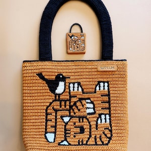 Op de afbeelding: Een gehaakte tote bag en bijpassende mini-tas in oranje, zwart en wit. De tote heeft een vogel en abstract ontwerp. De handvatten en afwerking zijn marineblauw. De mini-tas heeft een vergelijkbaar ontwerp. De merknaam "PEUM" staat op de tas.