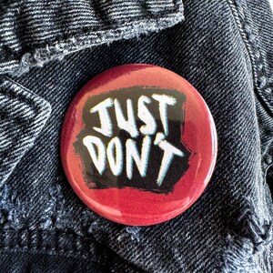 May include: A round, red button with the words "JUST DON'T" in white, outlined in blue, and set against a black background. The button is resting on a dark denim jacket.
