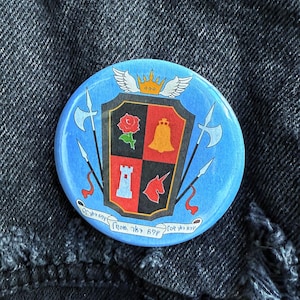 May include: A blue button with a crest featuring a rose, bell, tower, and unicorn. The crest is surrounded by two axes and a scroll with the text "For the love of the game from my heart".