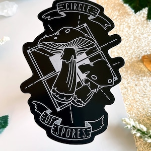 May include: Black and white sticker with a mushroom design. The sticker features two mushrooms with the text "Circle of Spores" in a banner.